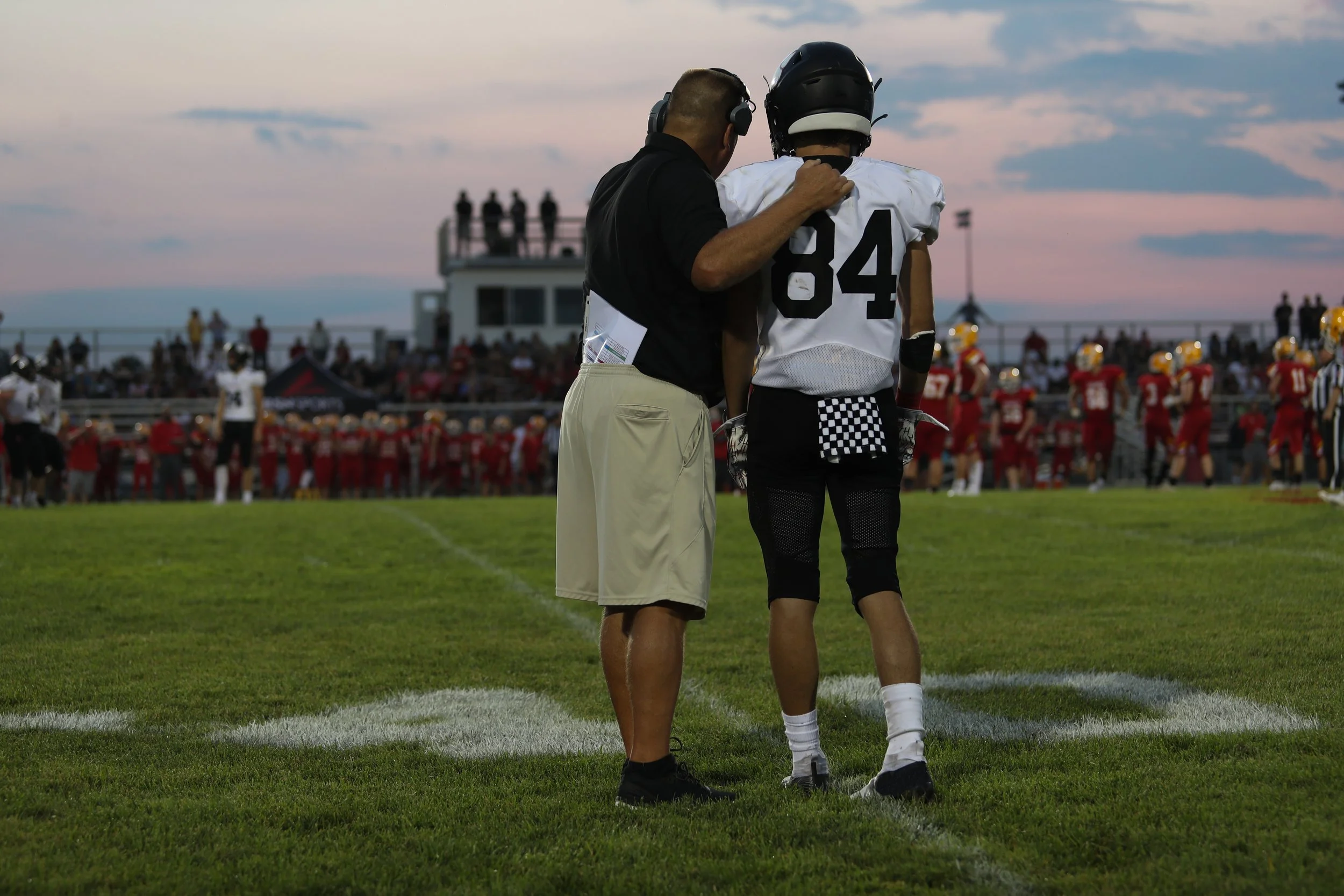 Football coach consoling quarterback on the field during a game at dusk, with players and spectators in the background.
