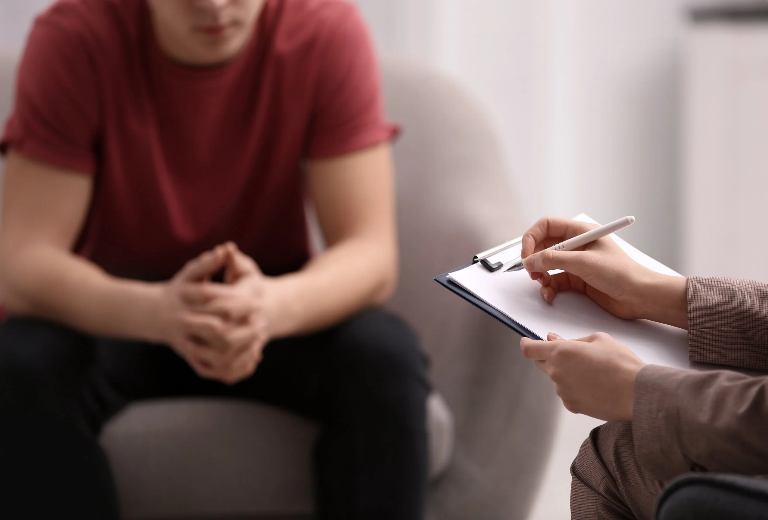 Person in casual clothing sitting on a couch with hands clasped, talking to counselor or therapist taking notes on a clipboard.
