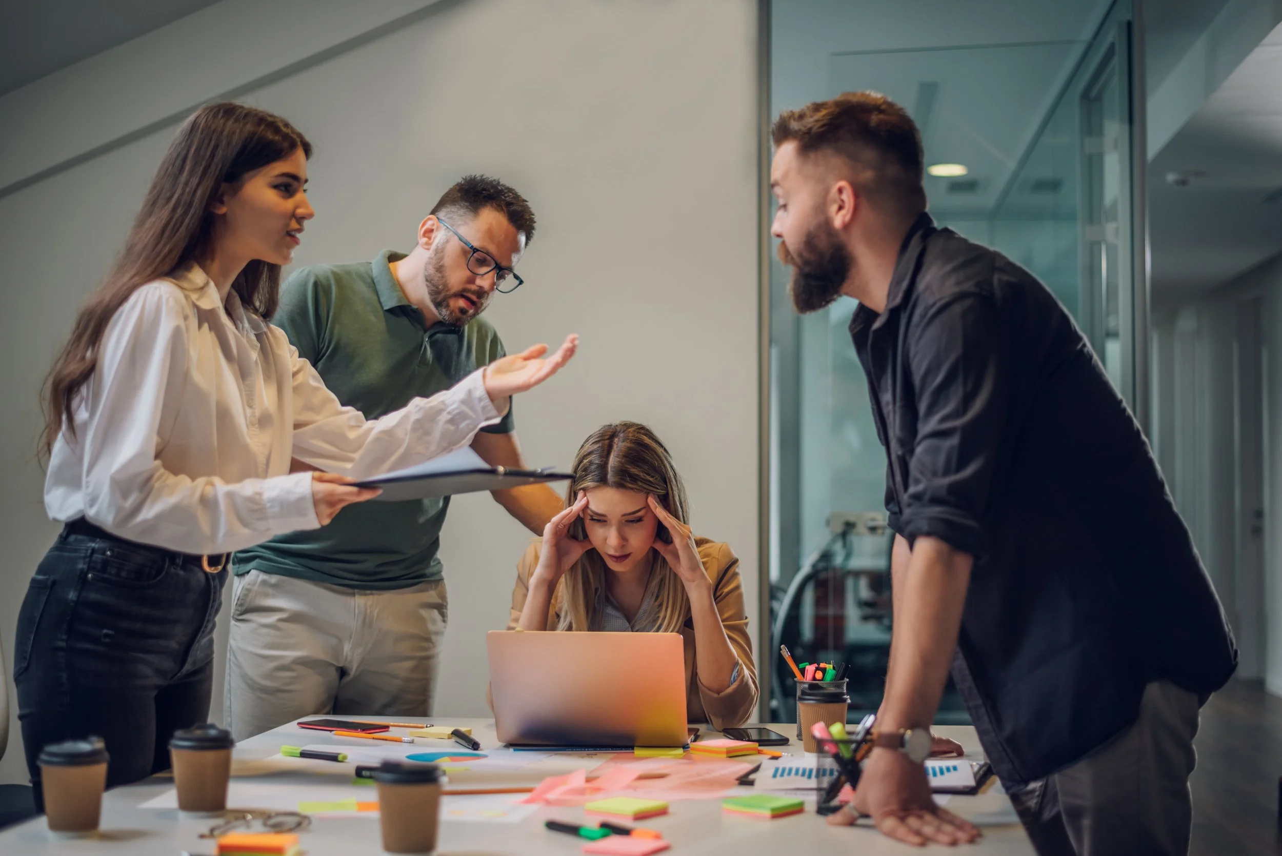 Team of five professionals having a stressful brainstorming session around a cluttered table with laptops, coffee cups, and colorful sticky notes.