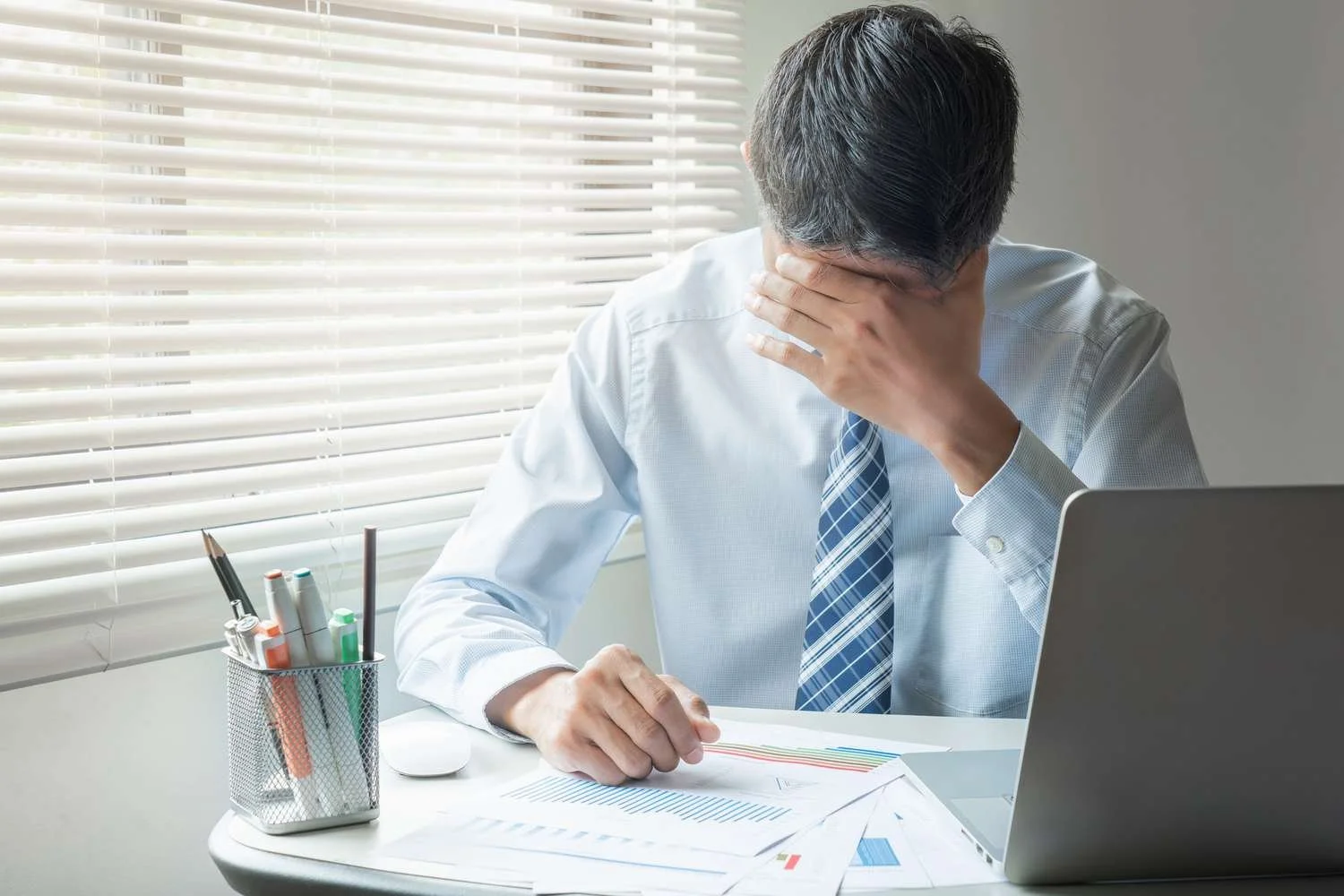 A man in business attire sitting at a desk, covering his face with his hand, with documents and a laptop in front of him, near a window with blinds.