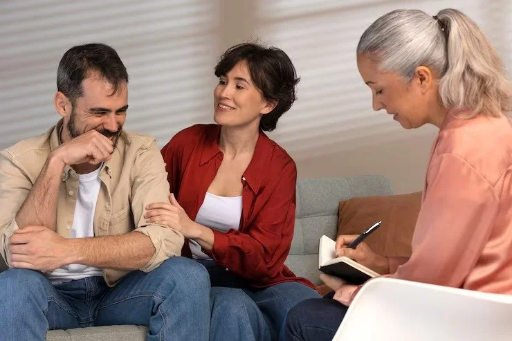 A woman in a pink top talking to a man and a woman in a red shirt, sitting on a couch, during a therapy session with a therapist taking notes.