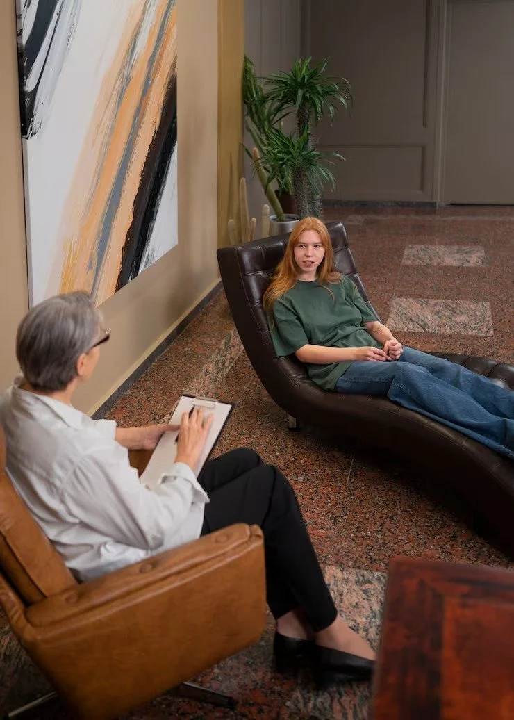 A young girl with red hair lying on a brown lounge chair, talking to a therapist who is sitting in a wooden chair with a notepad, in a therapy office decorated with an abstract painting and a potted plant.