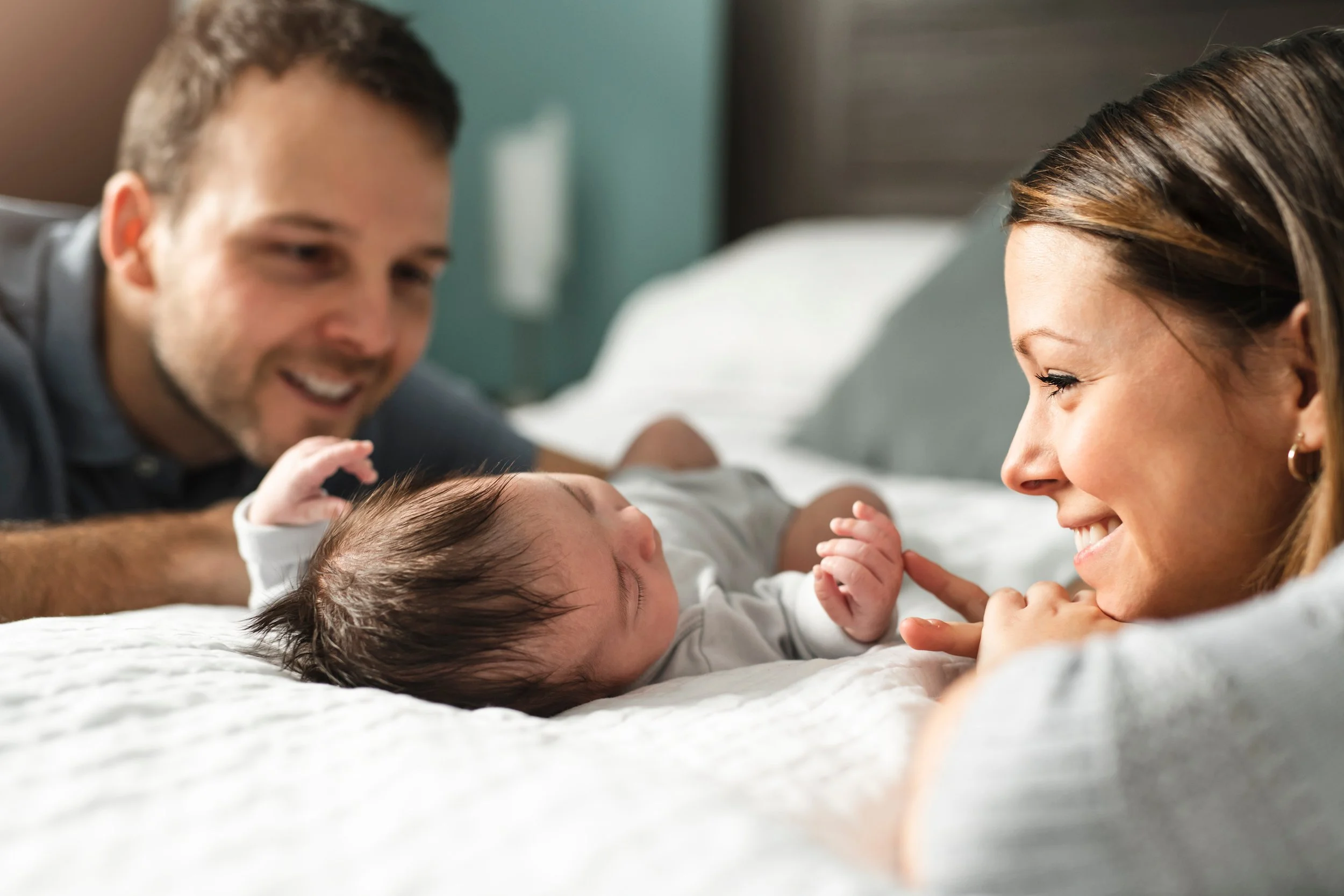 A happy family with a newborn baby lying on a bed. The father is in the background smiling, the mother is smiling and touching the baby's hand, and the baby is looking up with closed eyes.