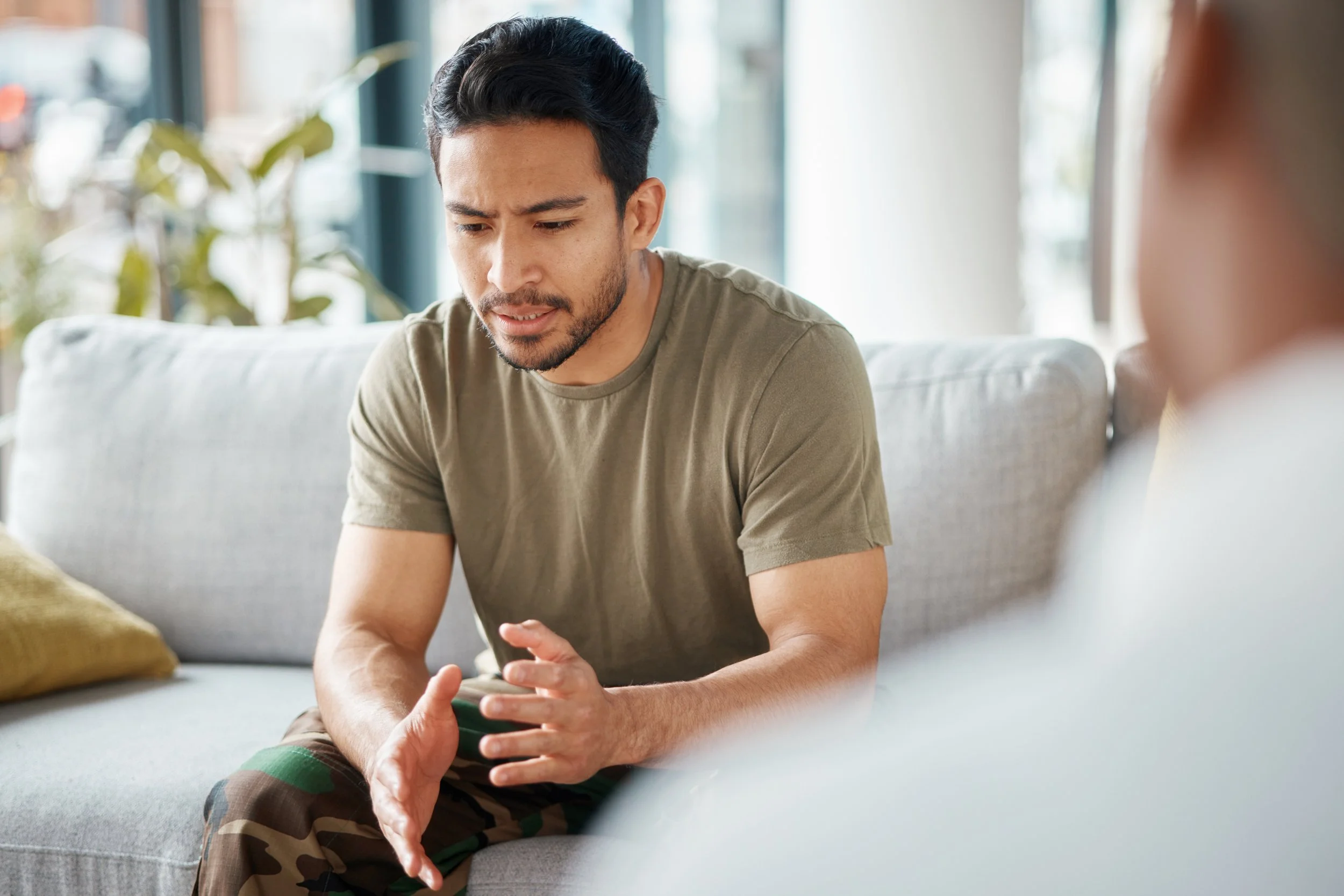 A man sitting on a light-colored sofa, appearing distressed, holding his hand and talking to someone off-camera.