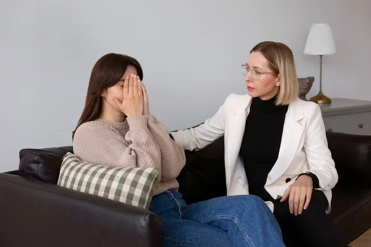 A woman in a beige sweater sits on a couch with her face covered by her hands, while another woman in a white blazer and black shirt comfort her with a gentle touch.
