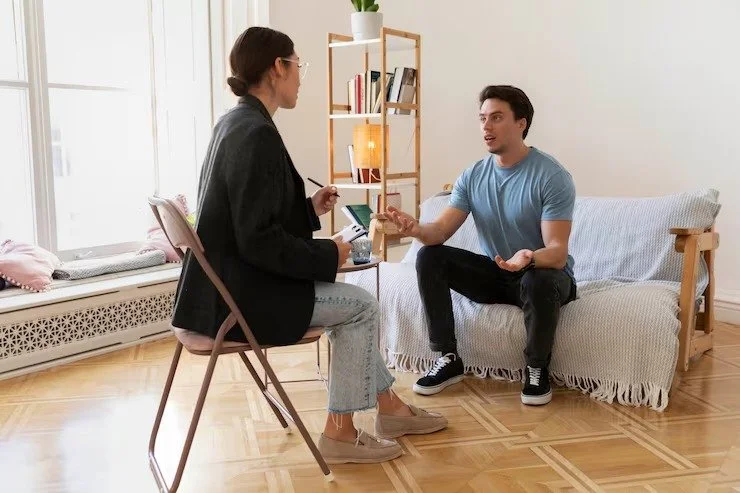 A woman and a man having a conversation in a living room, with the woman sitting on a pink chair and the man sitting on a sofa, gesturing with his hands.