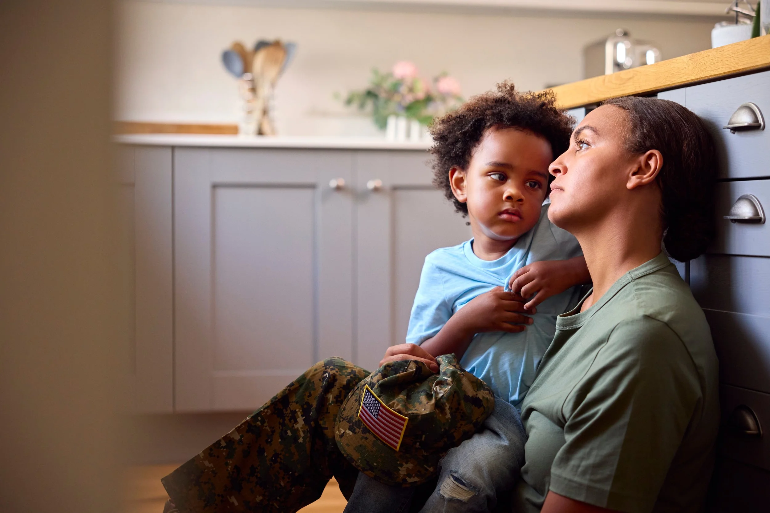 A woman in military fatigues sitting on the floor with a young boy in her lap, both leaning against a kitchen counter. The boy looks at her with a serious expression, and she gazes back at him, creating an emotional moment.