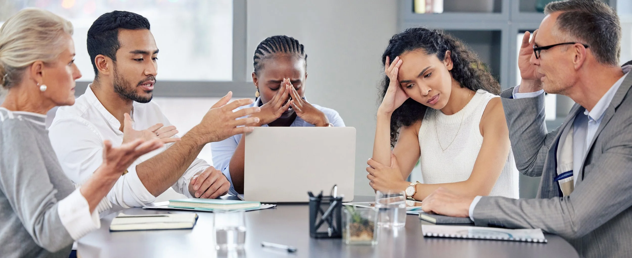 Group of five people sitting around a table, engaging in a serious discussion with stressed expressions, some with hands on their heads or face, in a professional setting.