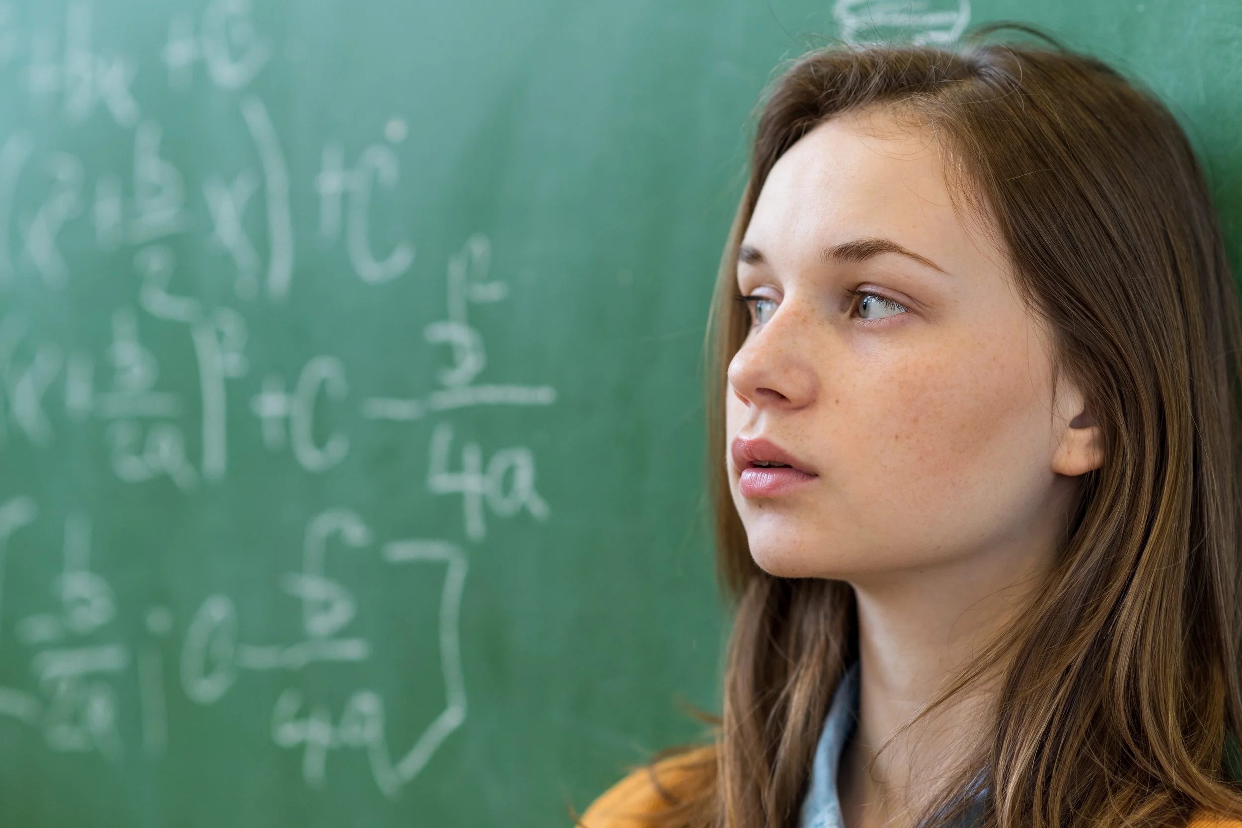 A young woman with brown hair and blue eyes standing in front of a green chalkboard with mathematical equations written on it.
