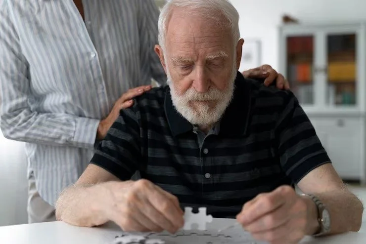 An elderly man with a white beard working on a jigsaw puzzle at a table, with a woman standing behind him offering support.