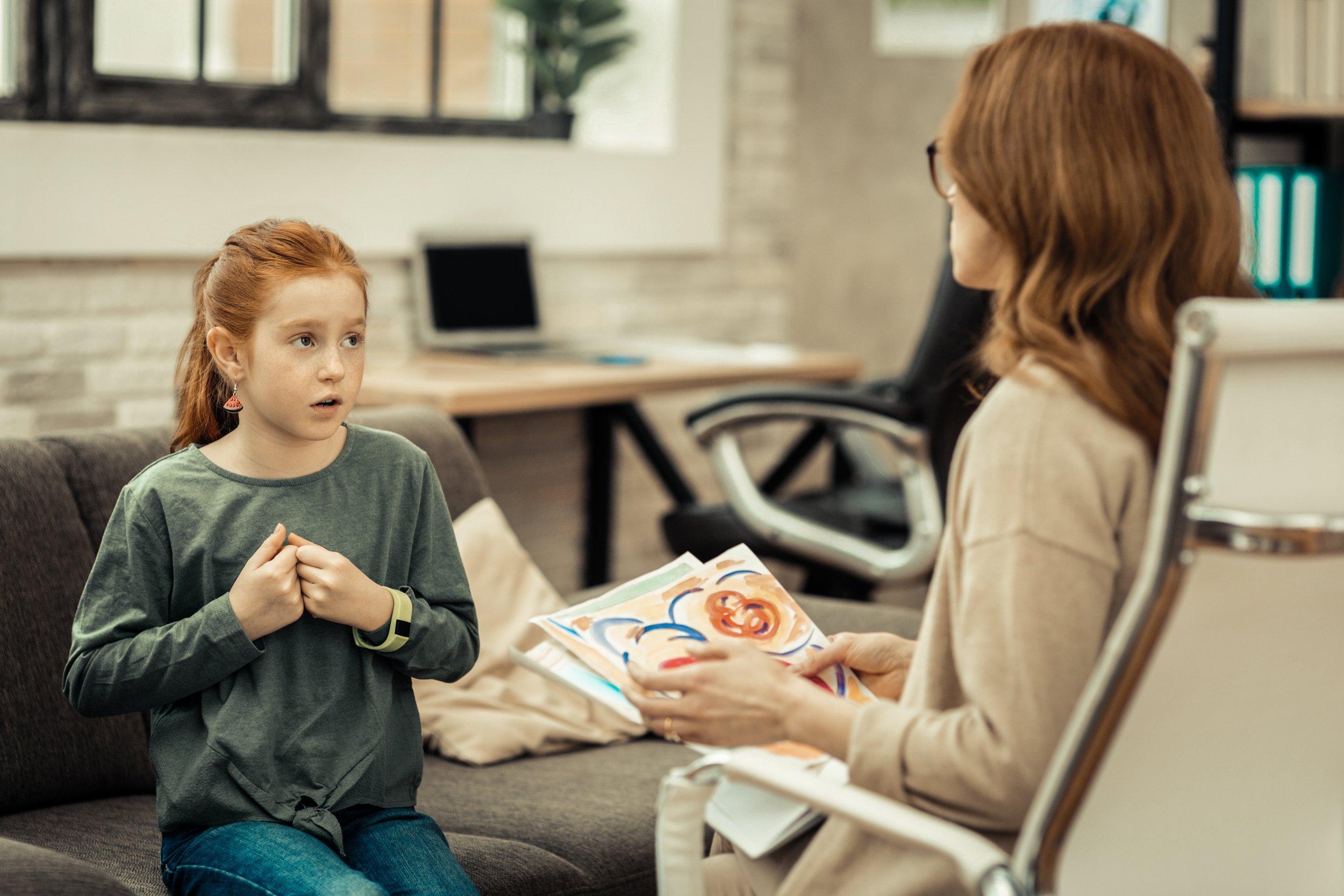 A young girl with red hair and a woman with brown hair in an office, sitting on a sofa and chair, engaged in a conversation. The woman holds colorful drawings or paintings, and the girl looks attentive and surprised.