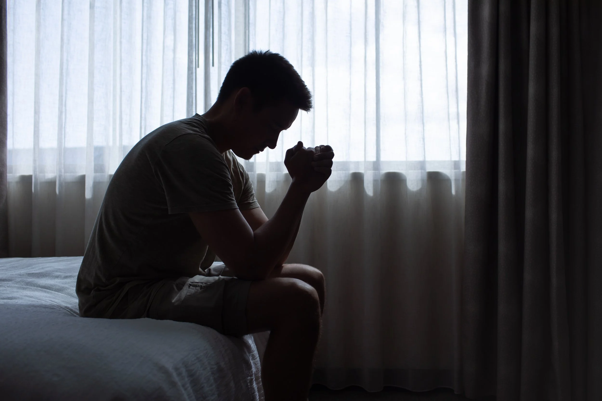 A man sitting on a bed with his head bowed and hands clasped together in prayer or contemplation, silhouetted against a window with curtains.