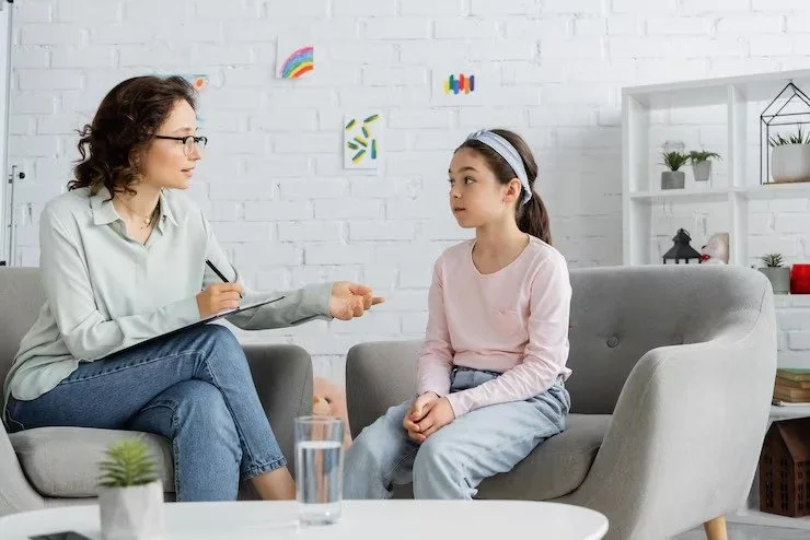 A woman and a young girl sitting on gray couches in a therapy or counseling session. The woman is holding a notepad and pen, talking to the girl in a comfortable, brightly decorated room.