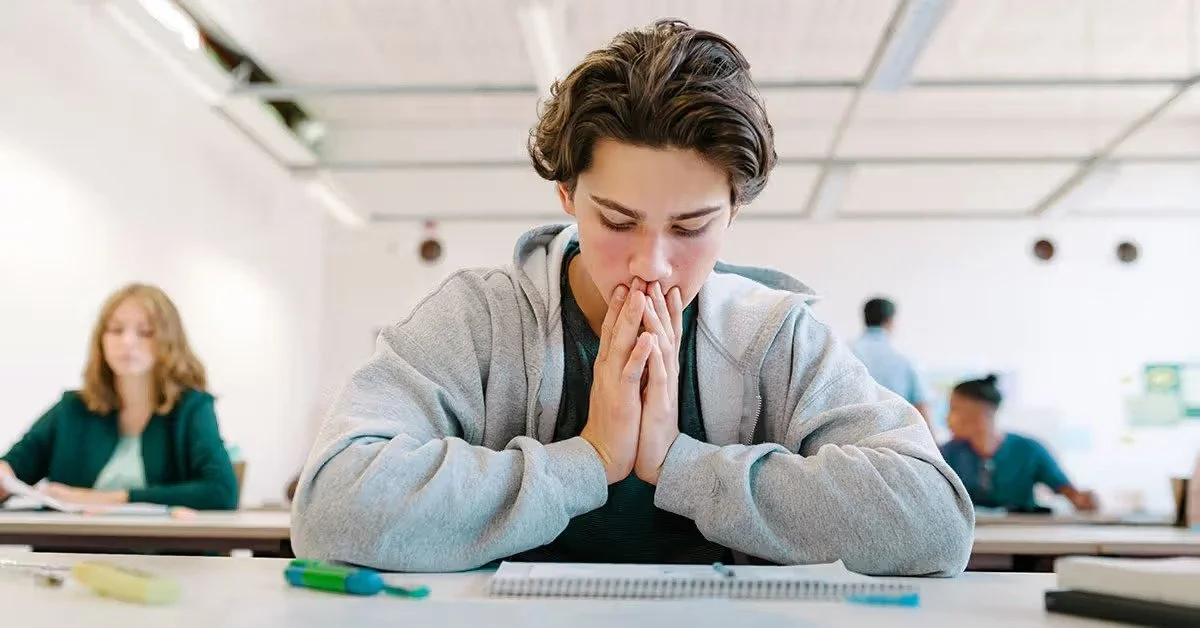 A young boy with dark hair sitting at a desk in a classroom, looking down with his hands clasped in front of his face, appearing worried or anxious.