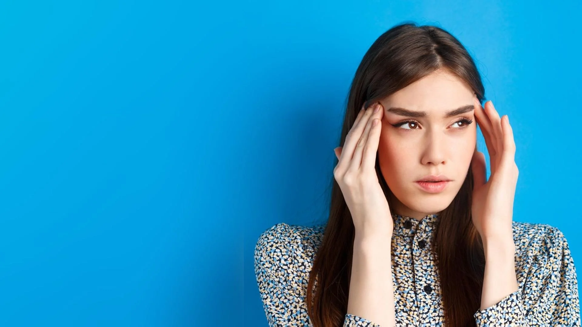Young woman with long dark hair holding her head with both hands, looking worried or stressed, standing against a blue background.