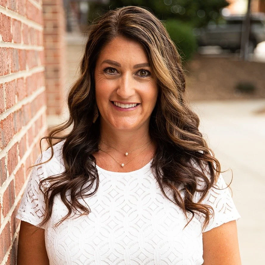 A woman with long, wavy brown hair and a friendly smile, wearing a white patterned top and layered necklaces, standing outdoors next to a brick wall.