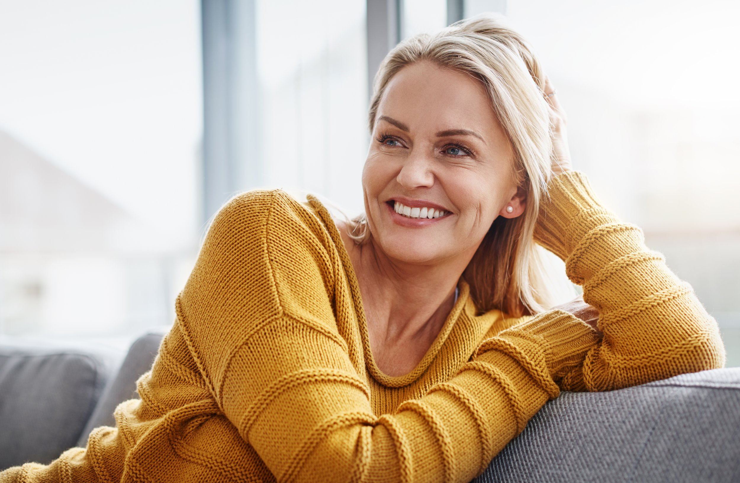 A woman with blonde hair smiling while sitting on a gray sofa in a brightly lit room, wearing a mustard yellow sweater.