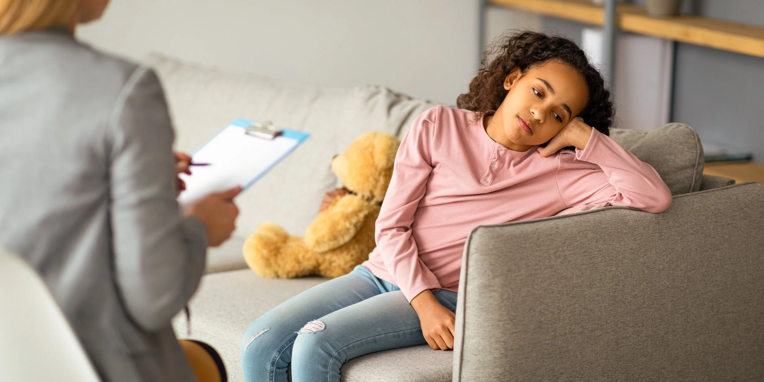 Young girl sitting on a couch looking tired or sad during a therapy session with a therapist taking notes.