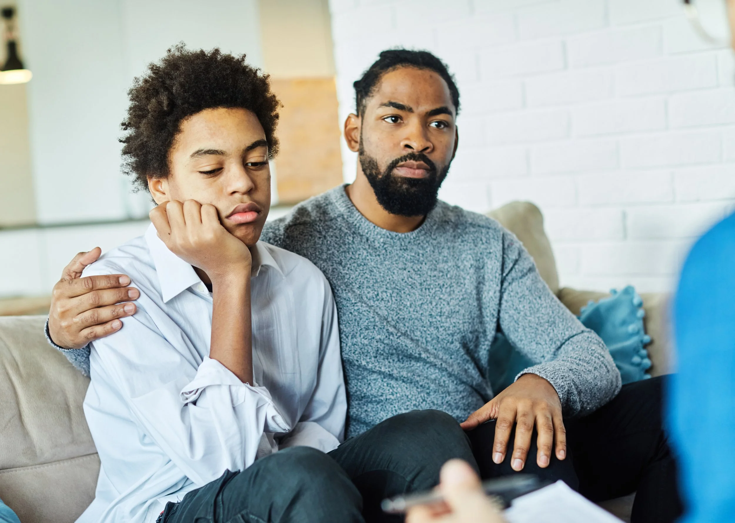 A worried teenage boy with curly hair sitting on a beige couch with a man who appears to be his father, on a white brick wall background.