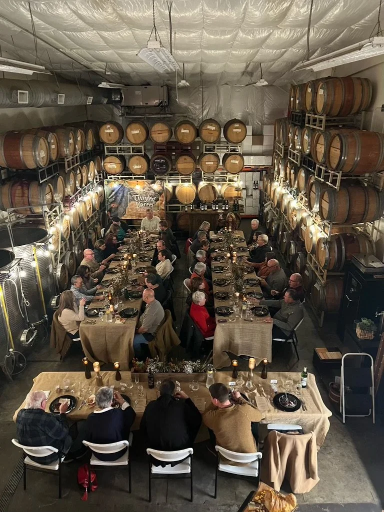 A large group of people dining at long tables in a brewery with rows of wine barrels on the walls.
