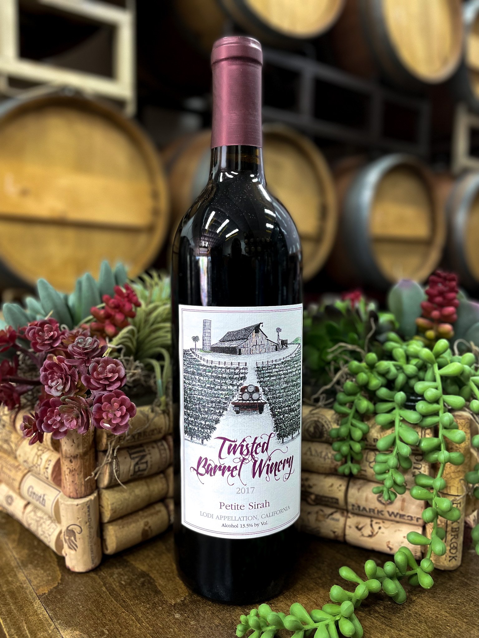 A bottle of twisted barrel winery petie sirah 2017 wine on a wooden surface surrounded by succulents and decorative wine corks, with wine barrels in the background.