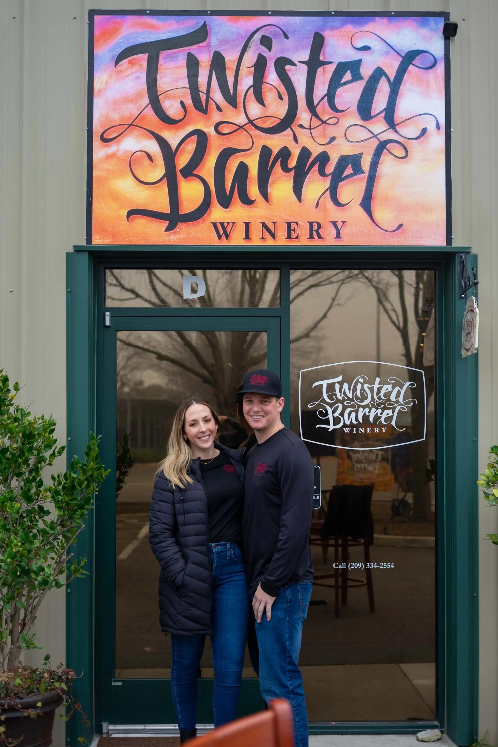 Two smiling people, a woman and a man, standing in front of the entrance to Twisted Barrel Winery. The woman has blonde hair and is wearing a dark jacket and jeans. The man is wearing a black jacket, blue jeans, and a black baseball cap. The winery sign is above them, and the glass door features the winery's logo.