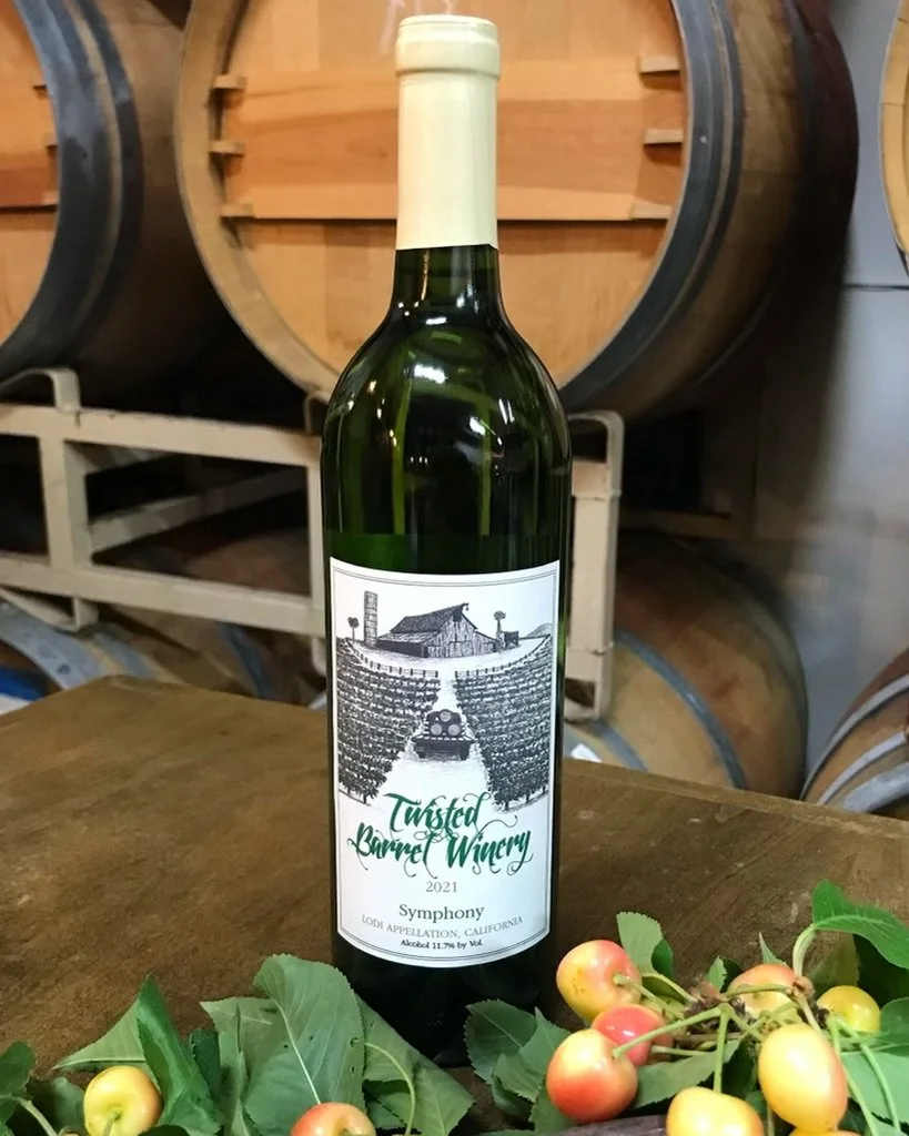 Bottle of Twisted Barrel Winery Symphony wine from Lodi, California, with a label showing a vineyard and winery scene, next to a small bunch of apples and leaves.