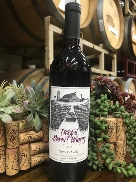A bottle of Twisted Barrel Winery Nero d'Avola wine from 2024 surrounded by decorative succulent plants and corks, with wine barrels in the background.