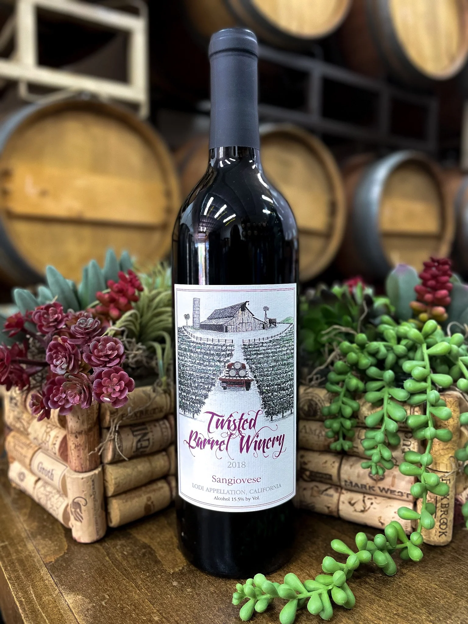 A bottle of Twisted Barrel Winery Sangiovese wine from 2018 in front of wine barrels and decorative plants.