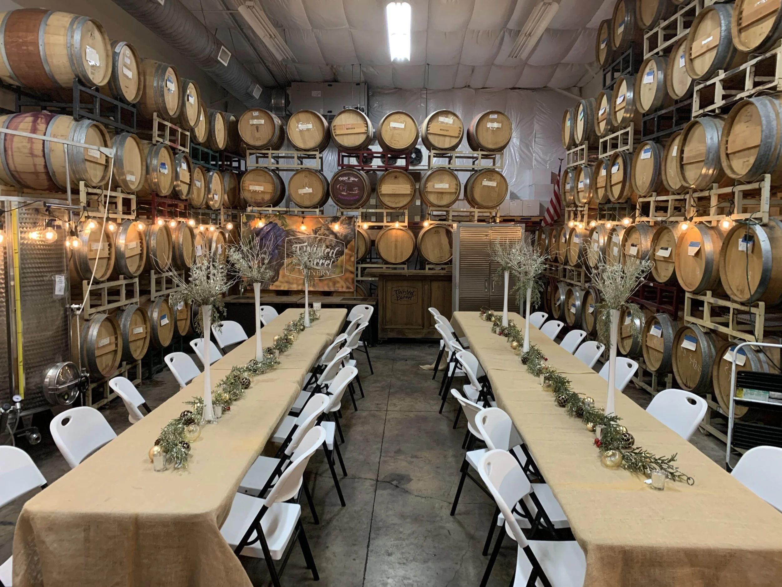 Long dining tables with white chairs, decorated with garlands, candles, and white vases with white branches. The room has walls lined with wine barrels and is set in a winery.