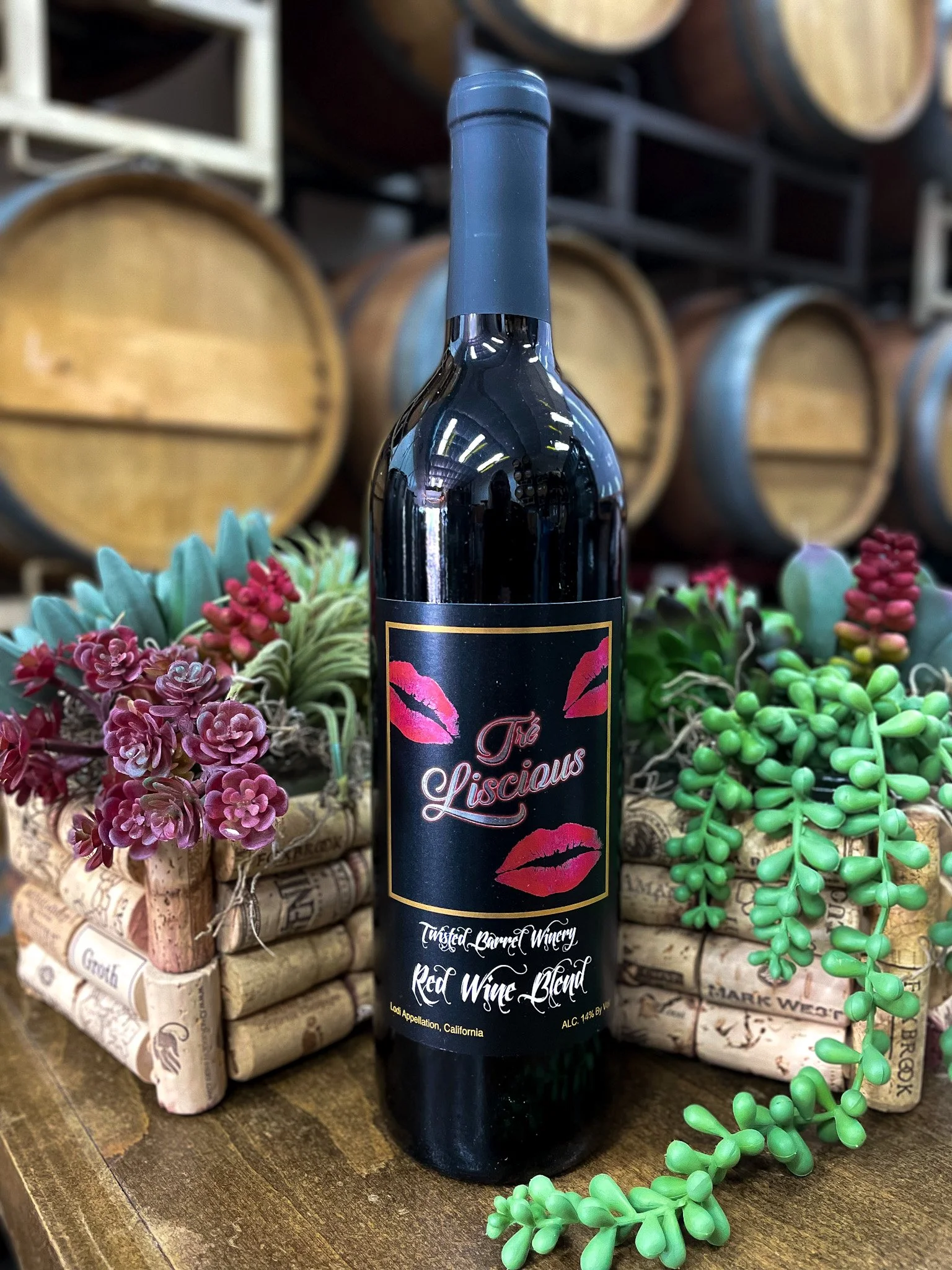 A bottle of red wine labeled "Tré Liscious" from Twisted Barrel Winery, placed on a wooden surface surrounded by small succulent plants and two decorative wine corks, with wine barrels in the background.