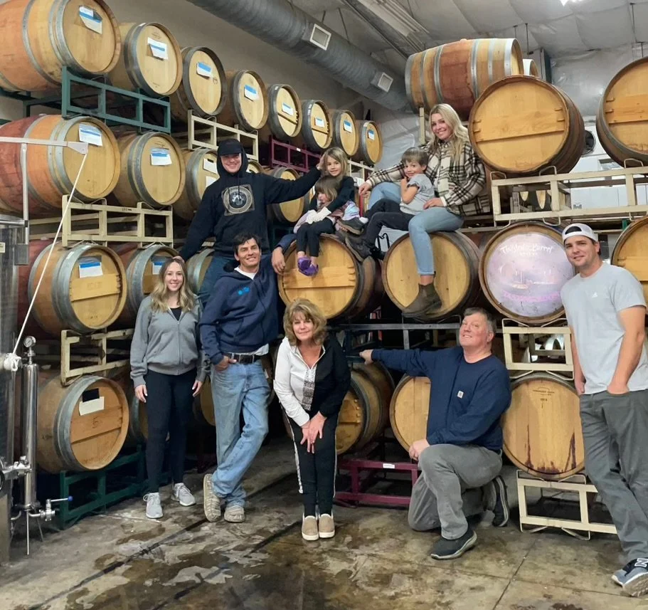 A group of nine people posing in a wine cellar surrounded by stacked wine barrels.