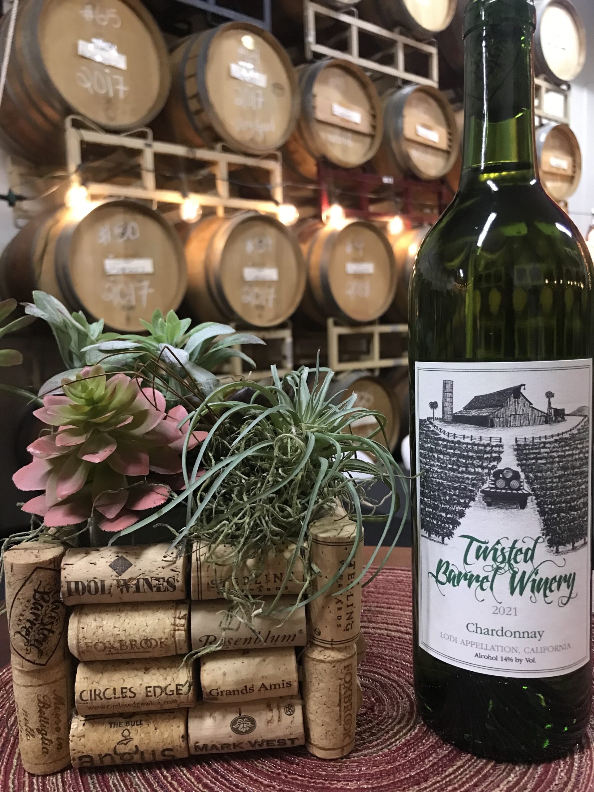 A bottle of Twisted Barrel Winery Chardonnay 2021 with wine barrels in the background, a small plant with succulents, and a decorative arrangement of wine corks on a table.