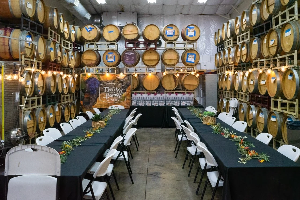 A winery event setup with long tables covered in black tablecloths, decorated with greenery and small flowers, surrounded by white chairs, and barrels stored on shelves on the walls, with a Twisted Barrel Winery banner in the background.