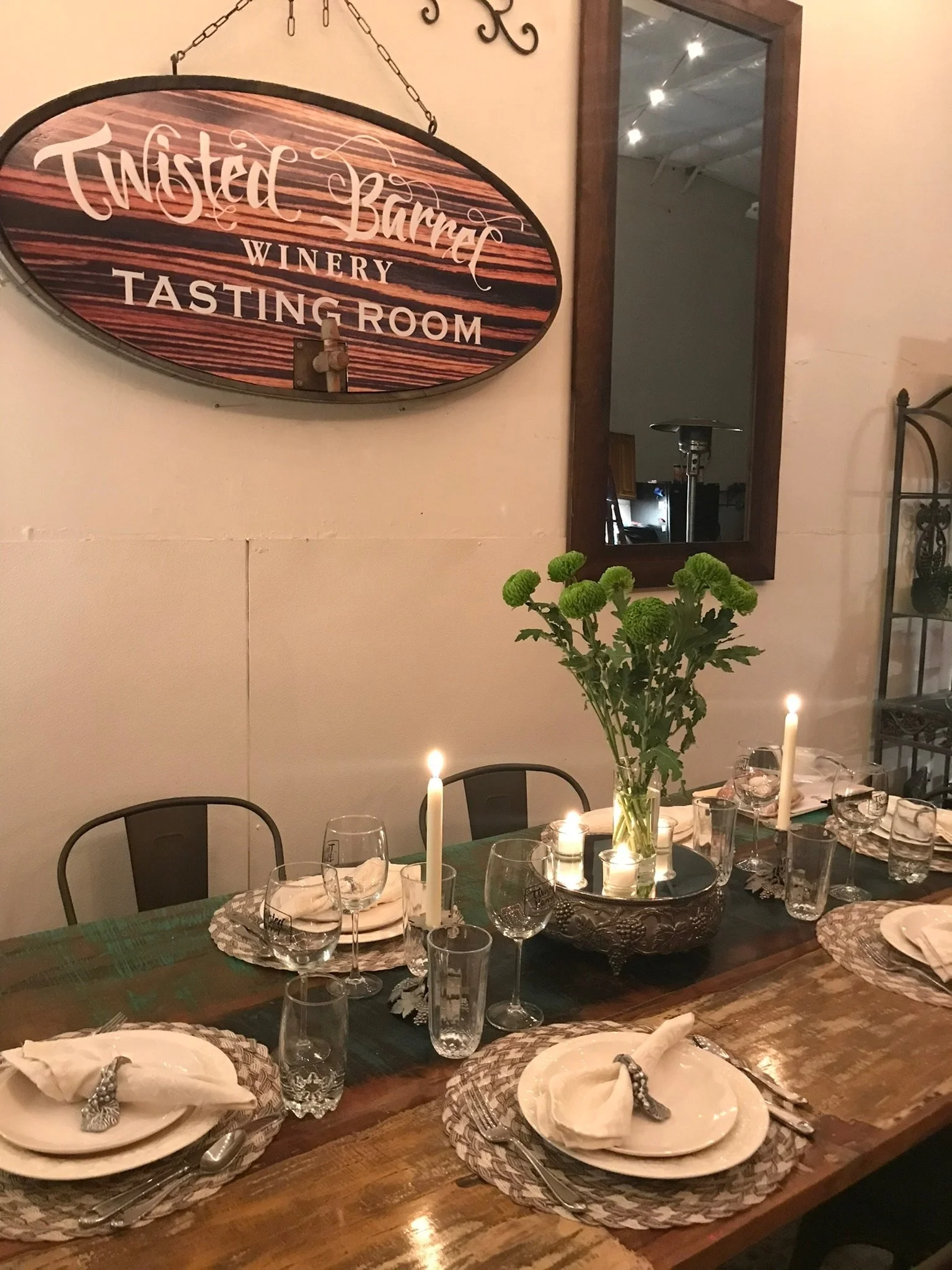 A dining table set with plates, napkins, silverware, glasses, and candles, with a vase of green flowers and a large mirror on the wall in the background. A sign reading 'Twisted Barrel Winery Tasting Room' is mounted on the wall.