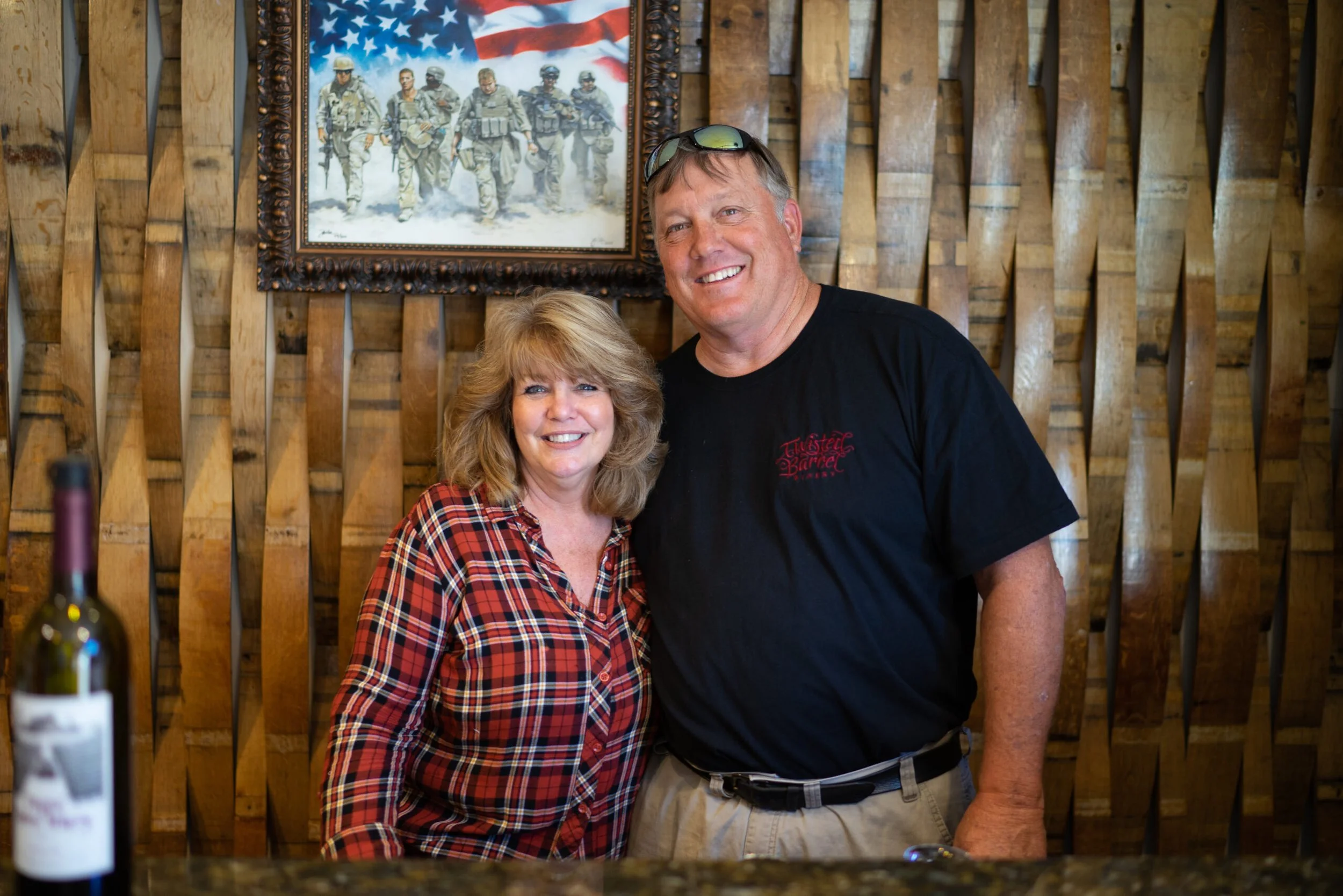 A smiling woman and man standing together indoors in front of a wooden wall with a framed picture of soldiers walking in snow. The woman is wearing a red and black checkered shirt, and the man is wearing a black T-shirt with red text and beige pants.