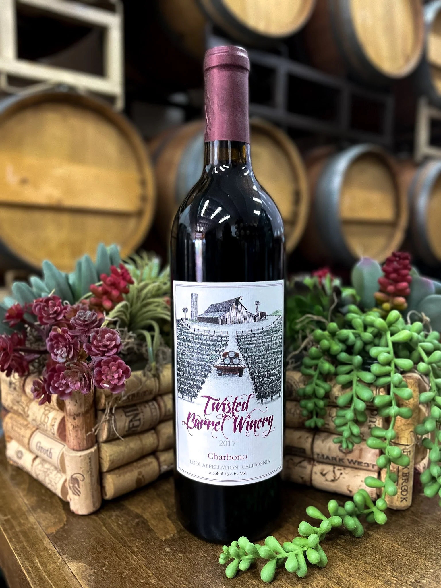A bottle of Twisted Barrel Winery red wine with a vintage vineyard scene on the label, placed on a wooden surface surrounded by succulents and wine barrels in the background.