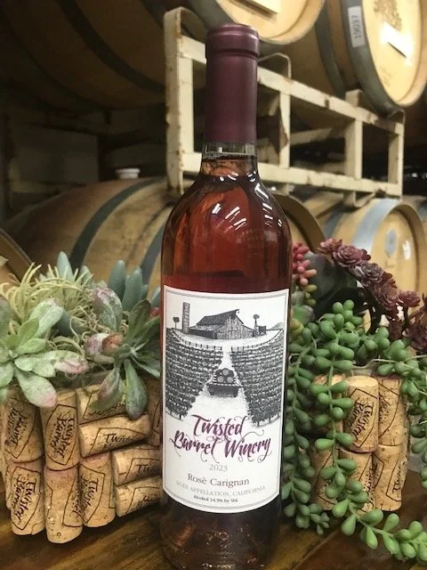 A bottle of Twisted Barrel Winery rosé wine, 2023 vintage, standing among grapevine corks and succulent plants with wine barrels in the background.