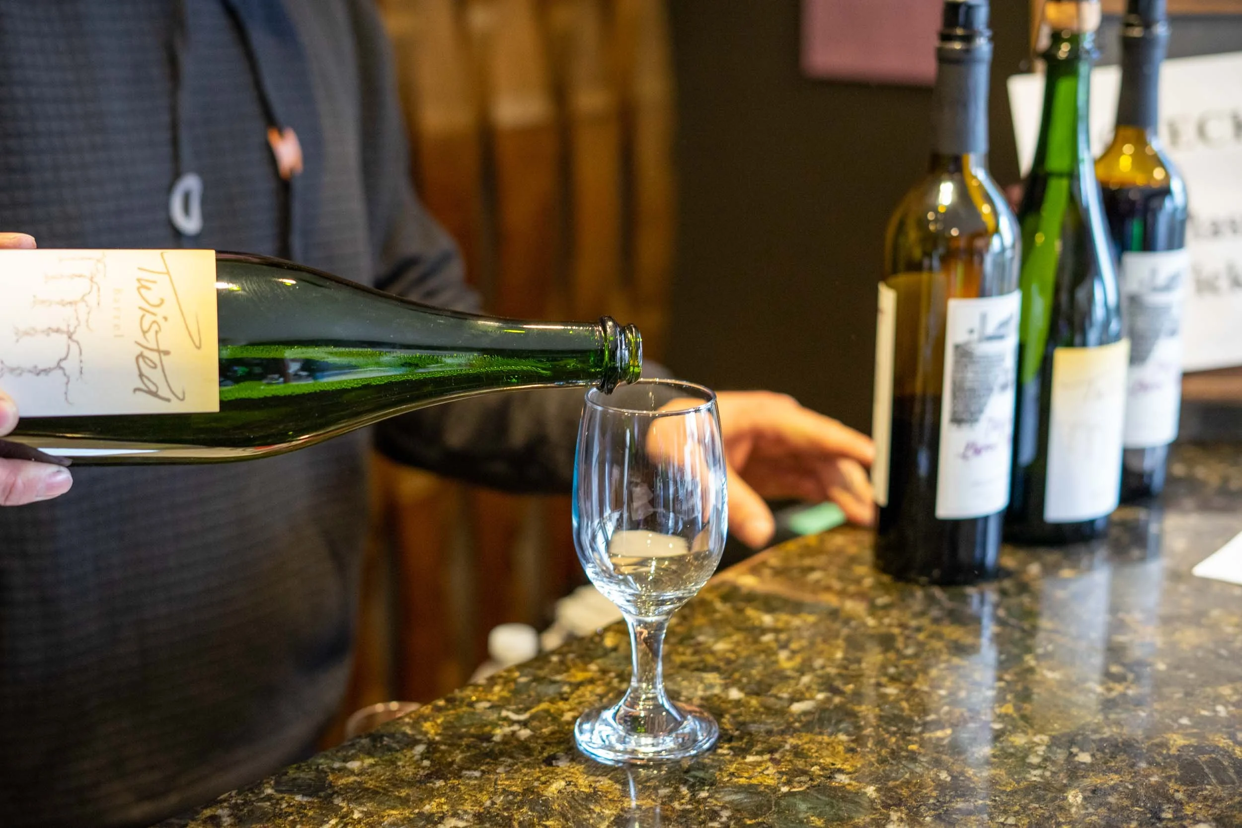 Person pouring white wine into a glass at a bar counter with wine bottles in the background.