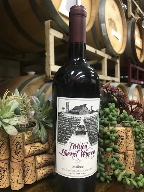 A bottle of red wine labeled "Twisted Barrel Winery 2022 Malbec" positioned among wine corks and succulents in a cellar or wine storage area.