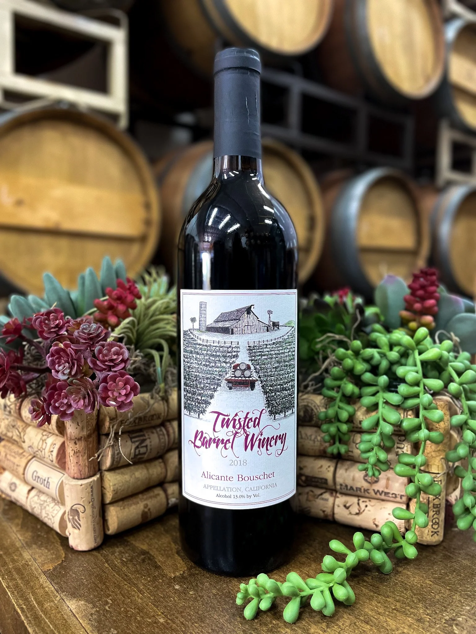 A bottle of Twisted Barrel Winery wine labeled 'Alicante Bouschet' from 2018, standing on a wooden surface surrounded by plants and decorative wine corks with an winery background of wine barrels.