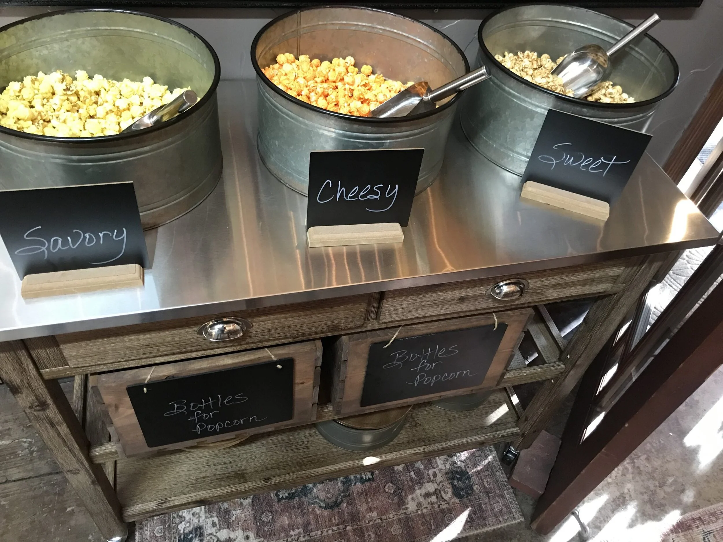 Three metal containers filled with different flavors of popcorn labeled 'Savory', 'Cheesy', and 'Sweet' on a counter, with a small chalkboard sign below reading 'Bottles for popcorn' and holding empty bottles.