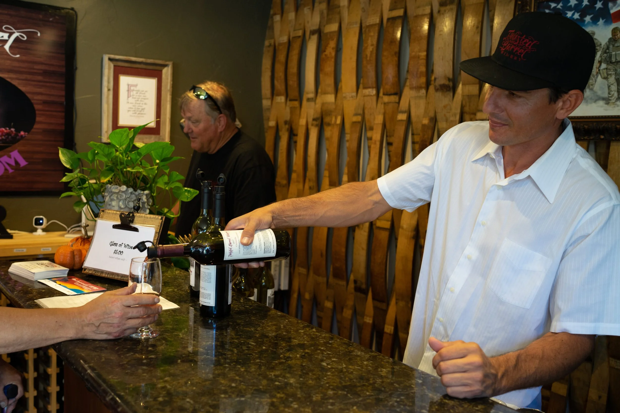 A man in a white shirt and black cap pours red wine into a glass at a wine bar, with another man in the background and a wooden wall behind them.