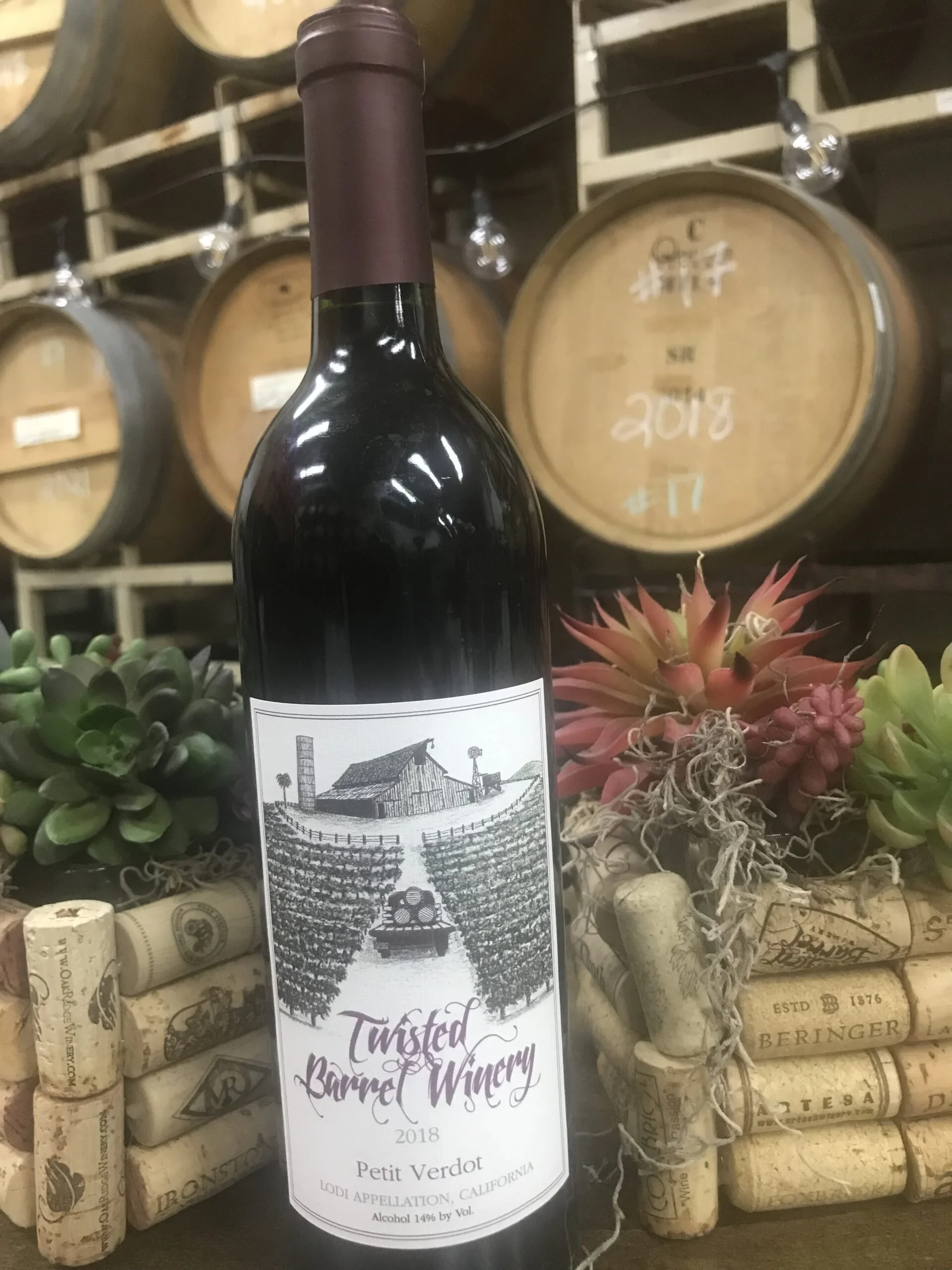 A bottle of Twisted Barrel Winery Petit Verdot wine from 2018, placed among wine corks, succulents, and drought-tolerant plants, with wine barrels in the background.