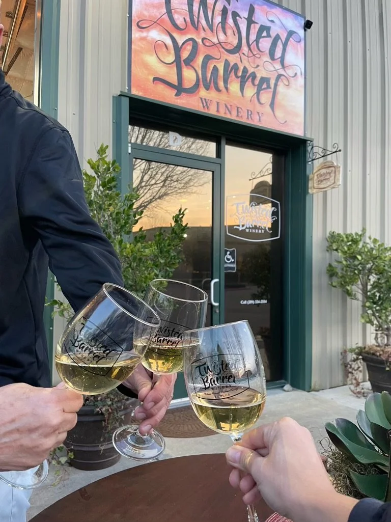 Three people clinking glasses of white wine outside Twisted Barrel Winery, with the storefront visible in the background, including the sign and plants.