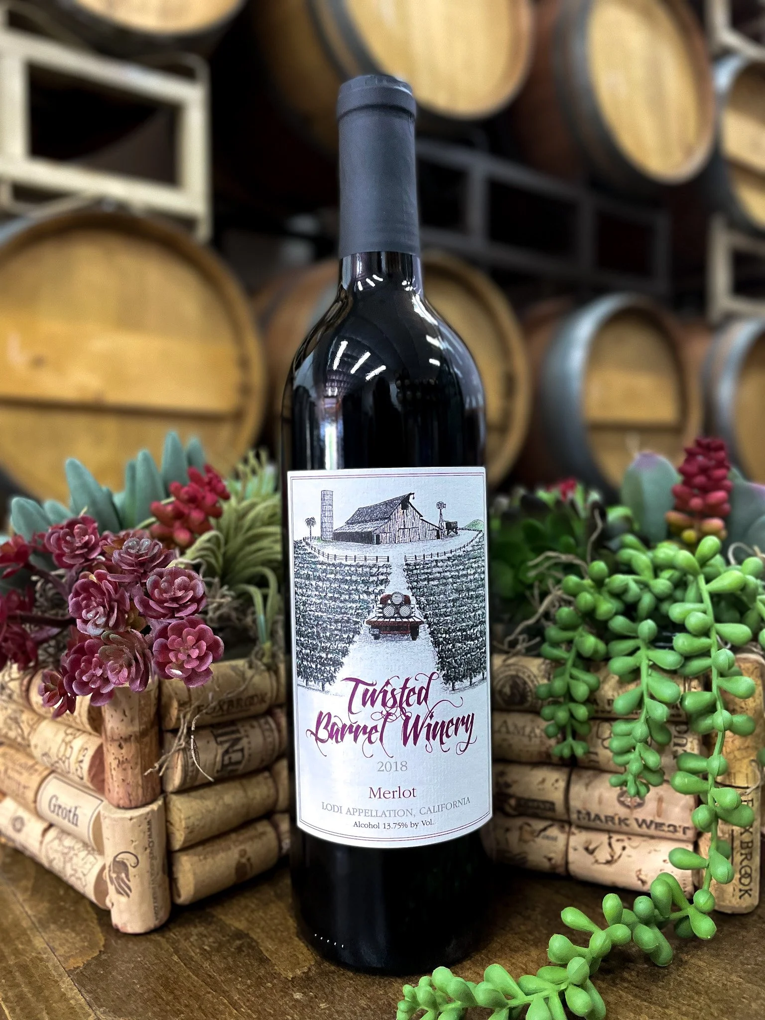 A bottle of Twisted Barrel Winery Merlot wine from 2018, placed on a wooden surface with decorative succulent plants and corks, with barrels in the background.