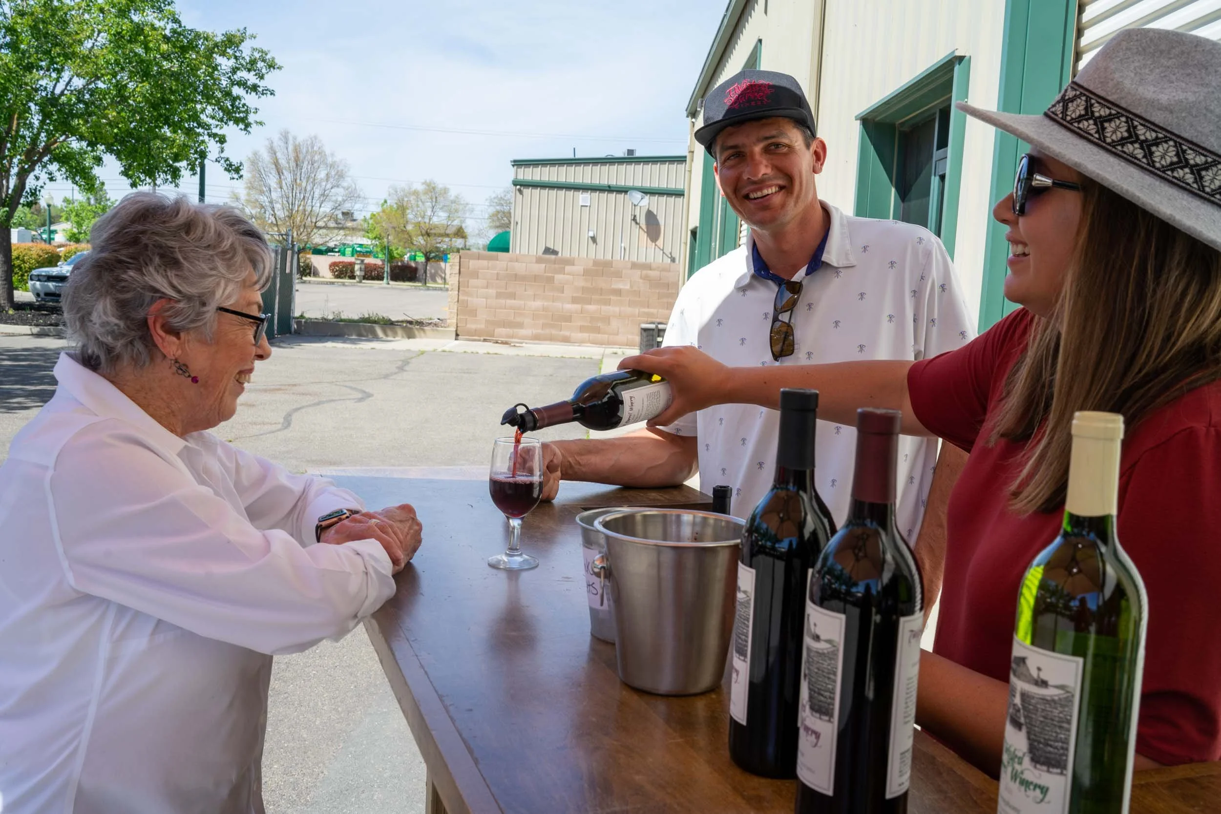 A woman receiving a glass of red wine from a server at an outdoor event, with two bottles of wine on the table.