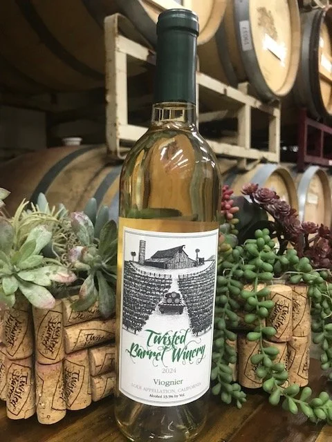 A bottle of Twisted Carrot Winery Viognier wine placed on a wooden surface surrounded by green and purple succulent plants, with wine barrels in the background.