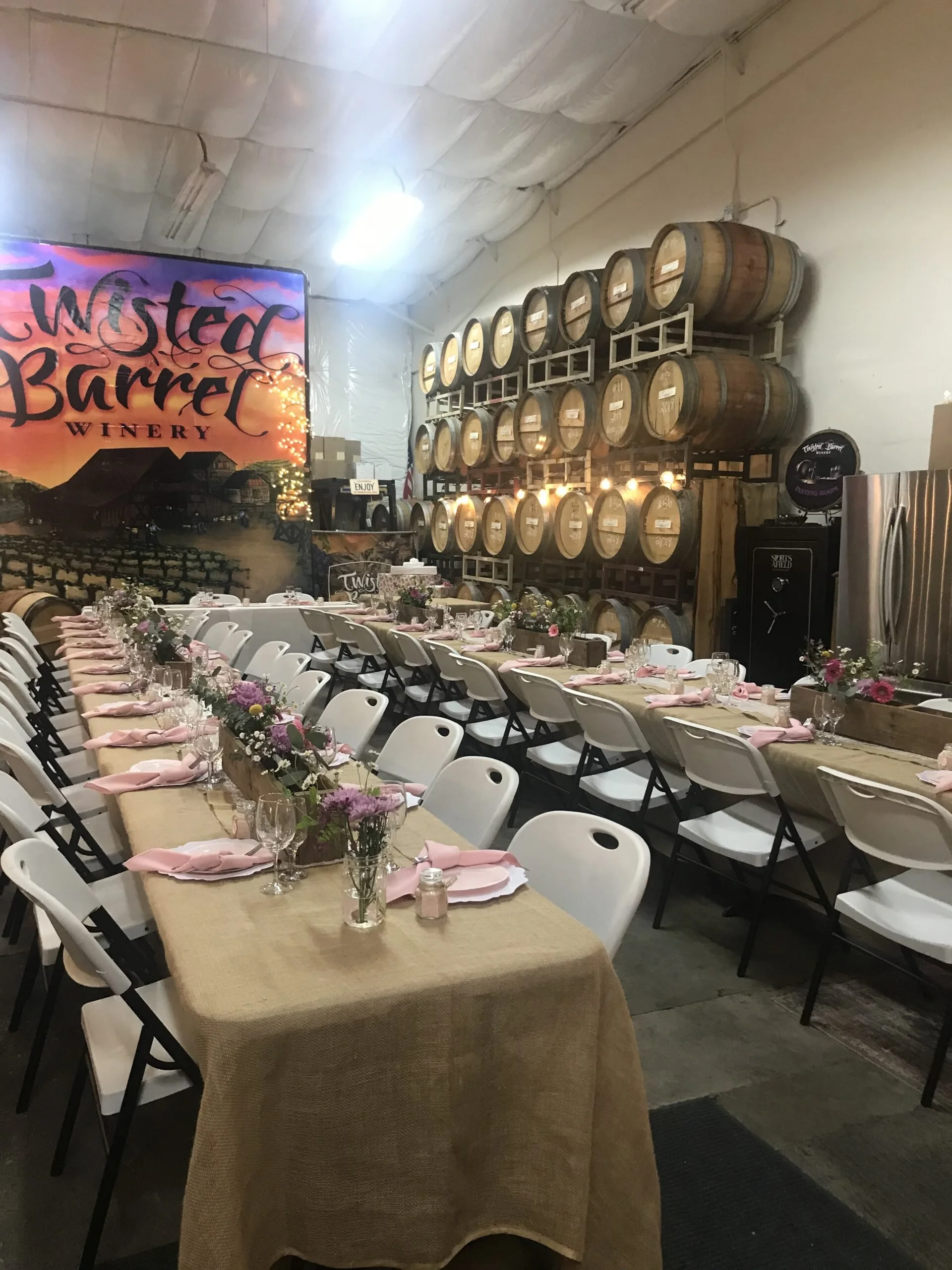 Set up for a celebration with long tables decorated with pink napkins, flowers, and glassware inside a winery with barrels and a Twilight Barrel Winery sign in the background.