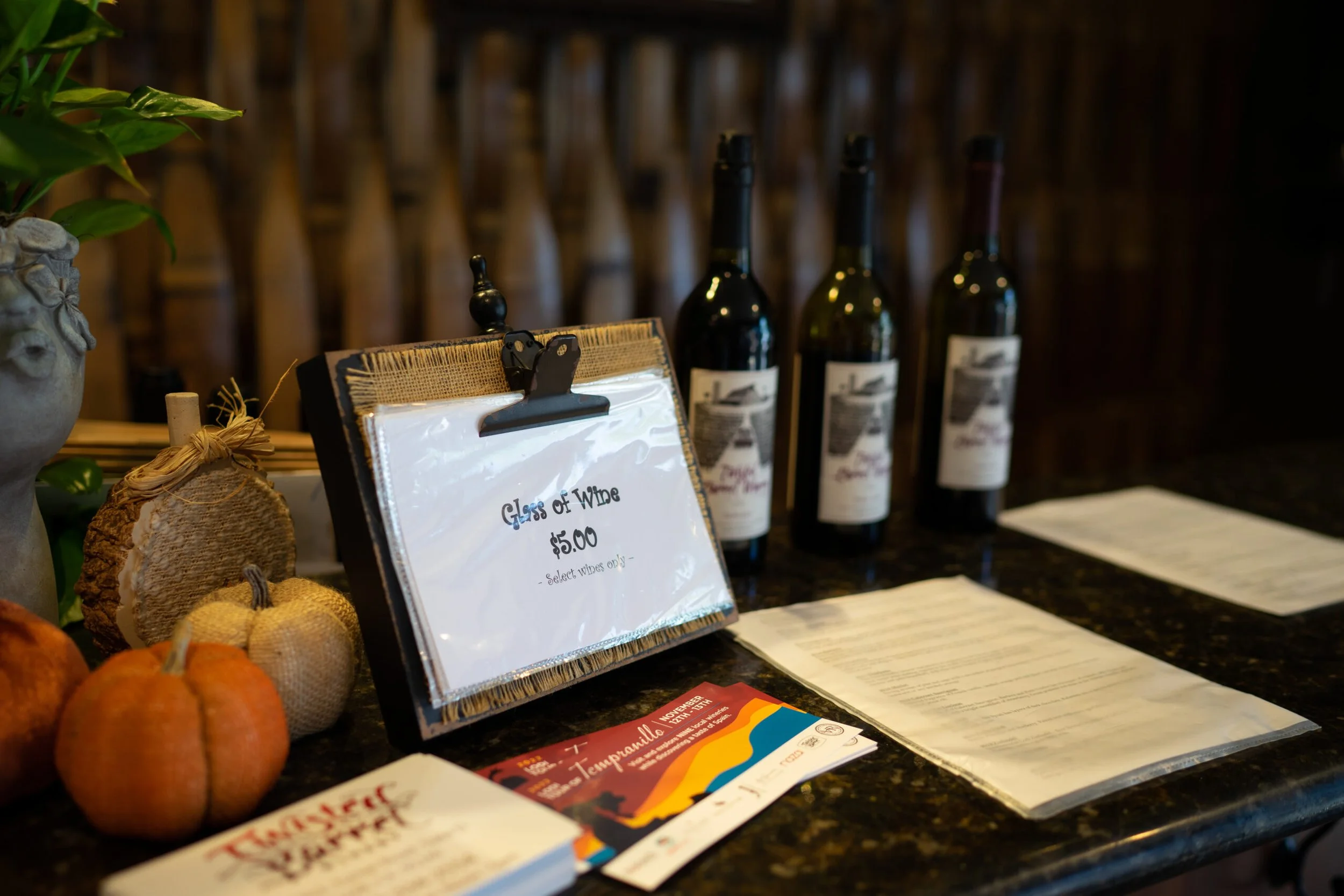 A display of wine bottles, pumpkins, and informational papers on a dark countertop with a rustic wooden background.